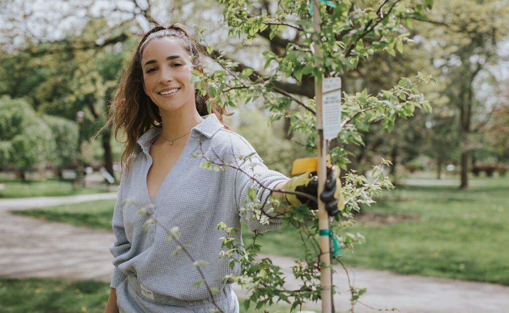 Gold medalist Aly Raisman wearing gray jumper holding the tip of a tree for earth month.