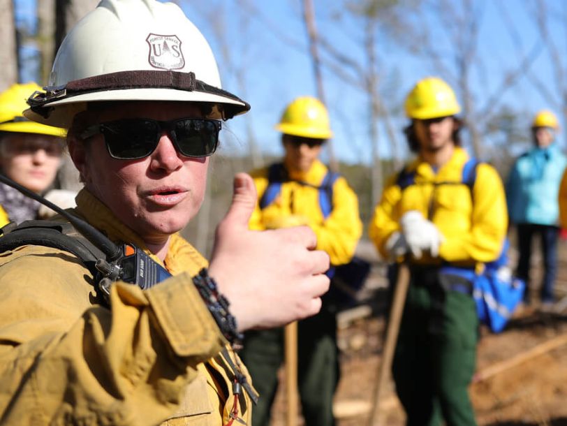 A woman in a uniform wearing a hard hat and sunglasses gives the thumbs up.
