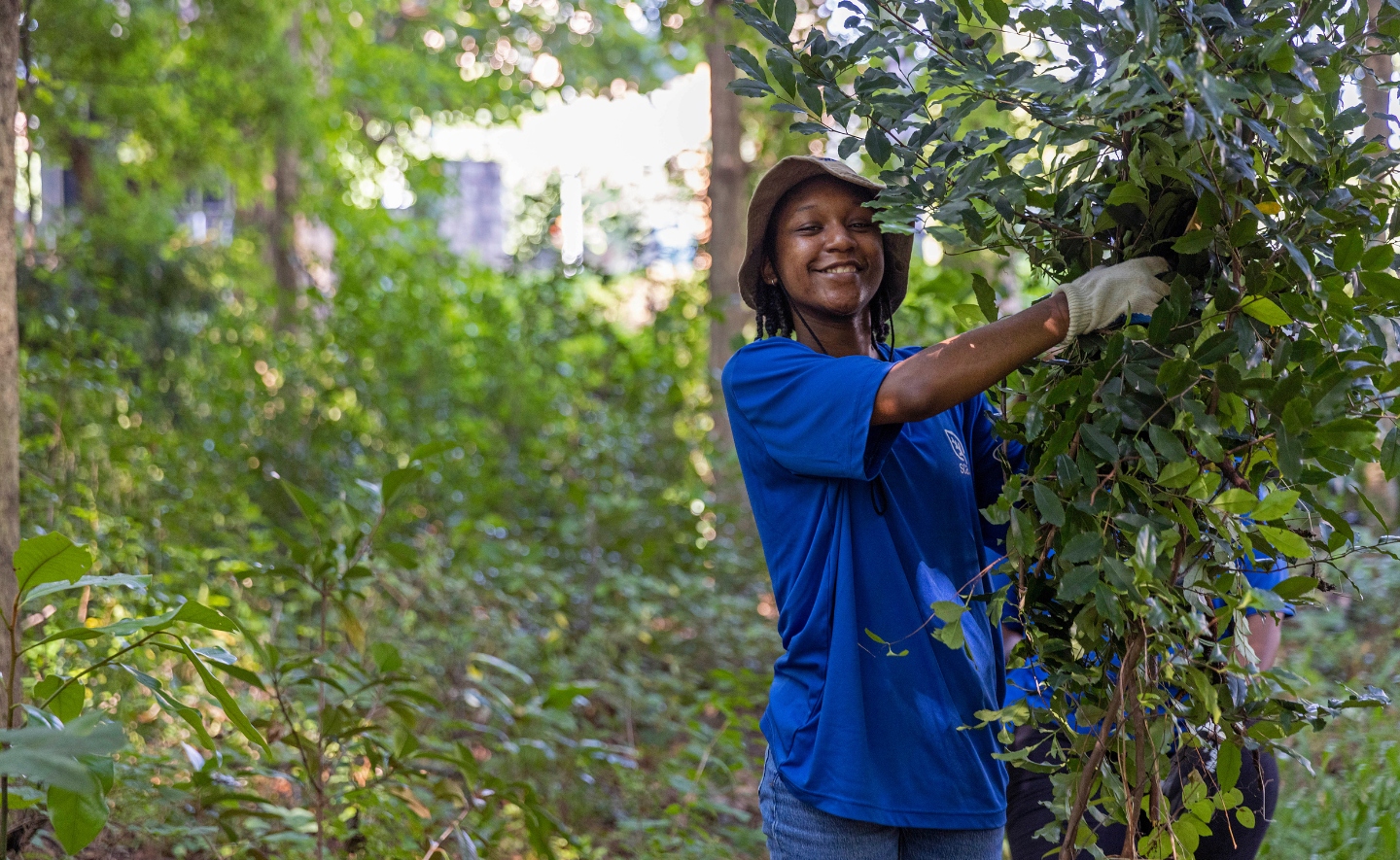 Atlanta Urban Green member wearing a blue t-shirt, tan bucket hat and white gloves holding cut tree branches.