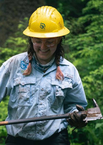 A person standing in the forest wearing a hard hat and holding an axe.