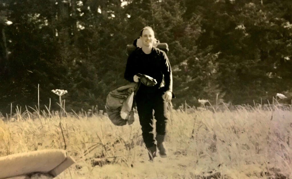 Vintage photo of SCA Alum Tommy Sheridan walking through a field carrying camping gear