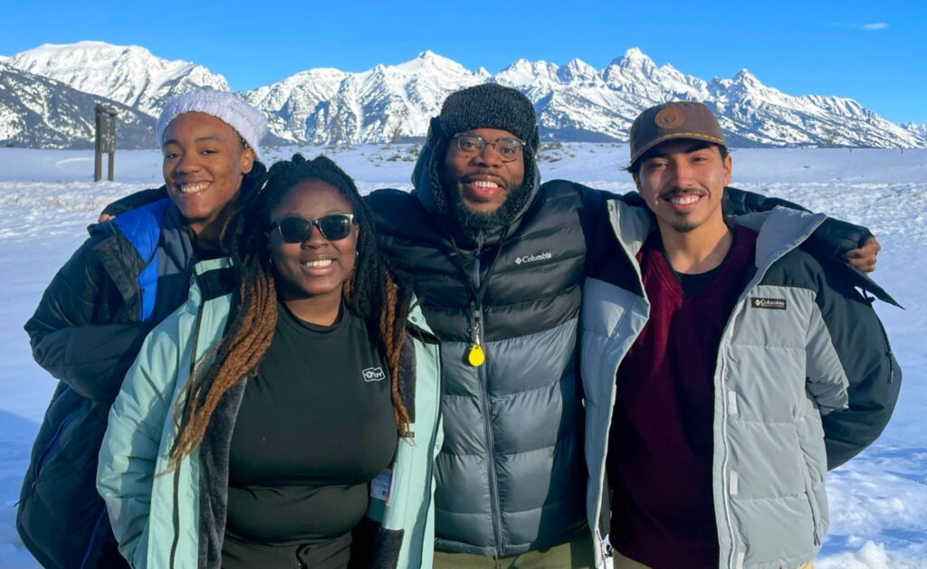 Four people stand together in heavy winter gear. Snow-covered mountain peaks are visible behind them.