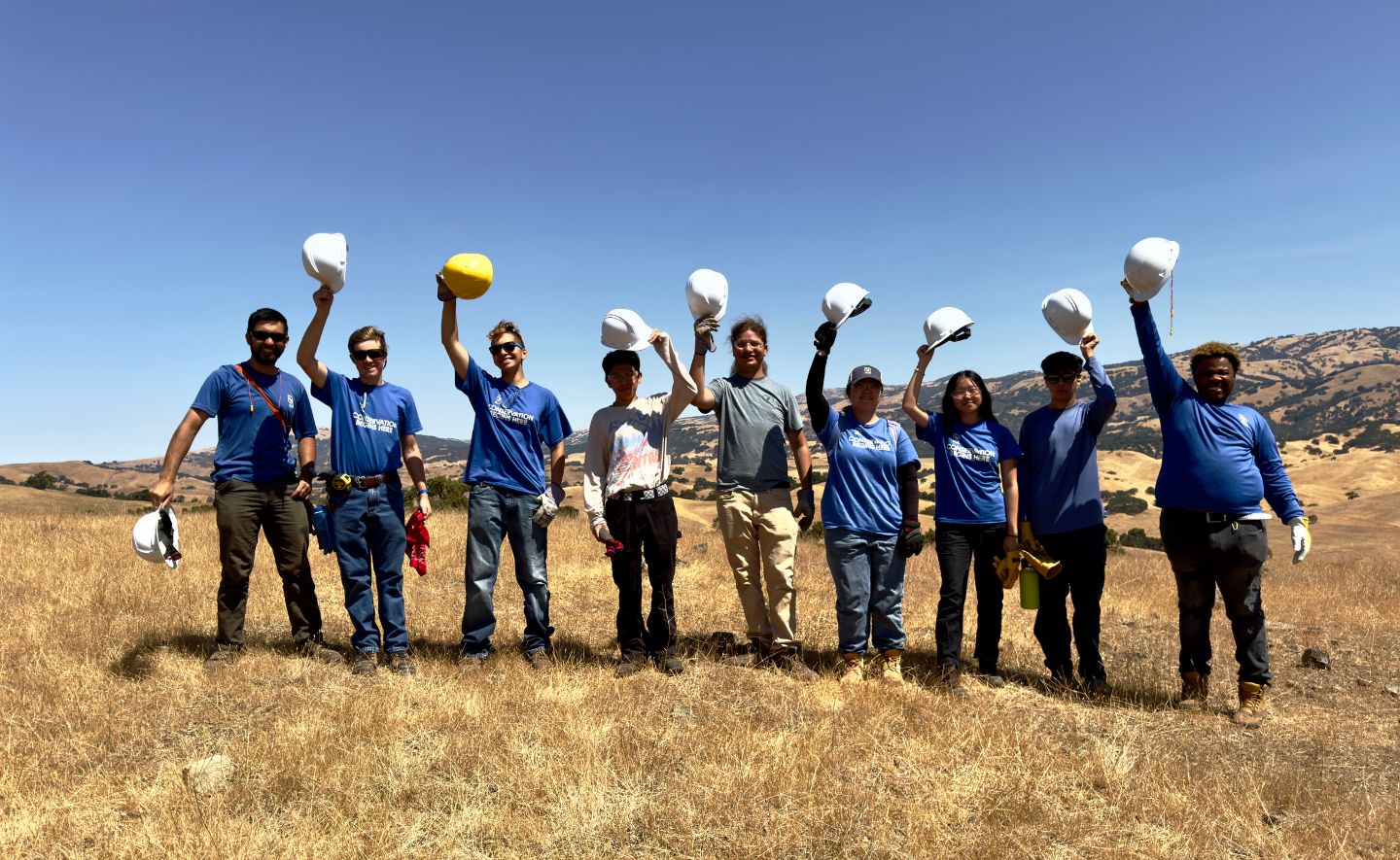 Bay Area Peninsula Climate Resiliency Crew is standing in a field holding up hard hats.