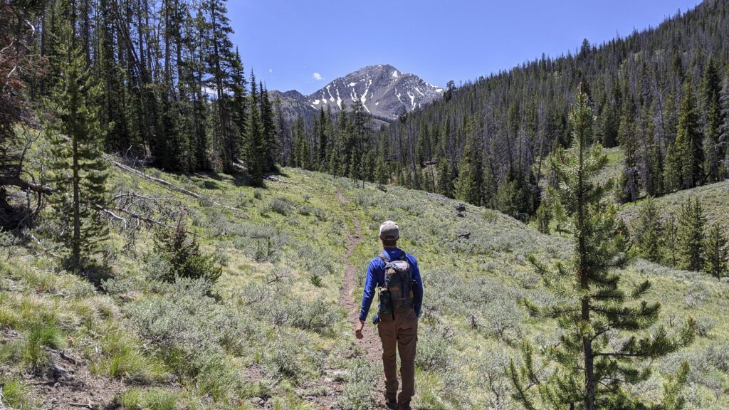 A lone hiker in a bright blue SCA shirt strides forward along a mountain path toward a peak striped with snow.