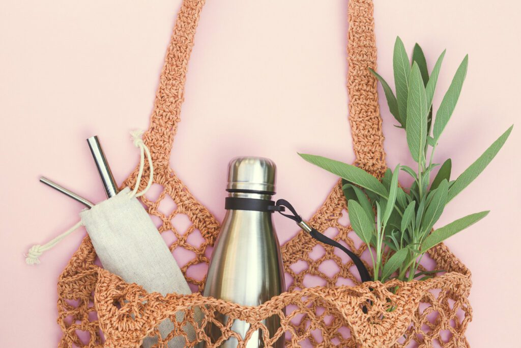 Reusable straws, a metal water bottle, and a plant in a woven reusable bag.