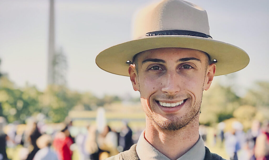 Kyle Yarusso stands wearing his Park Ranger uniform. The Washington Monument is visible in the blurred background.