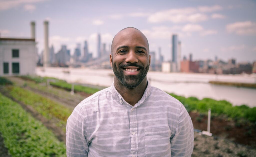 CJ Goulding stands outside in front of a blurred city background with neat rows of plants in the foreground.