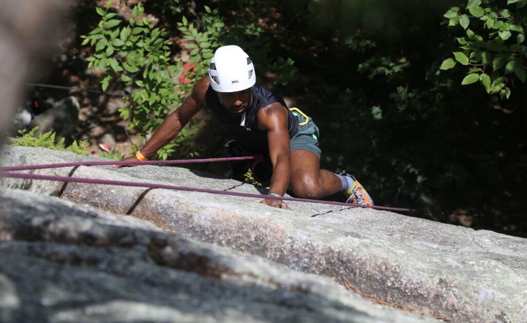 CJ Goulding wears a helmet and climbing gear as he scales a vertical rock face. Greenery is visible behind him.