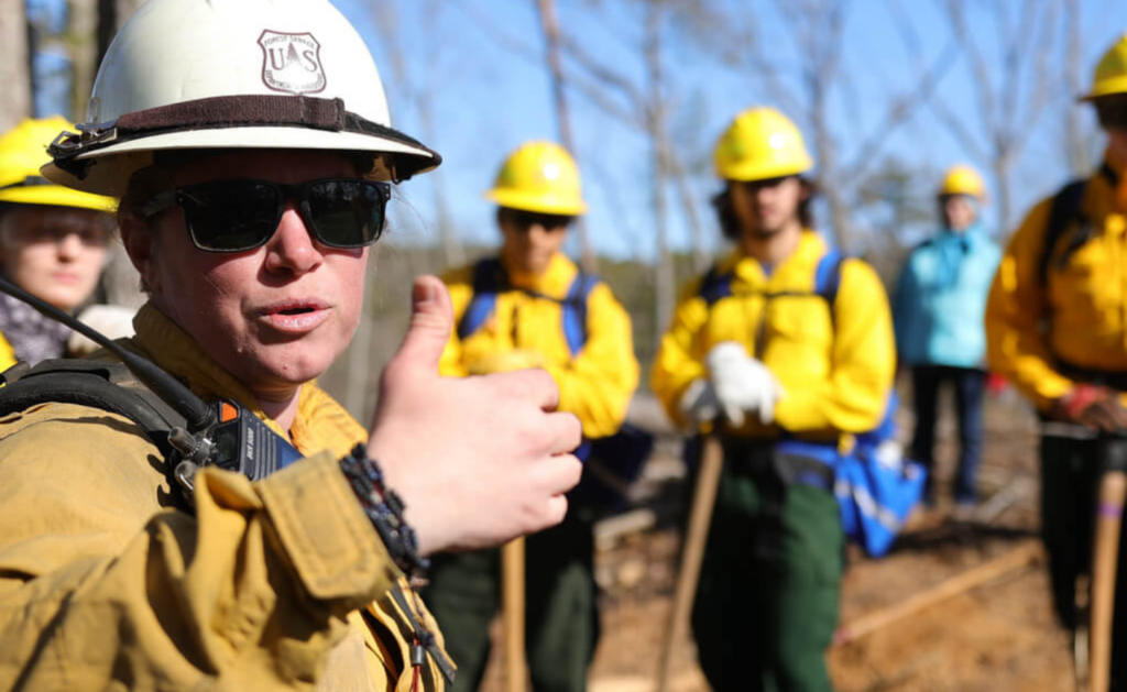 A woman in a uniform wearing a hard hat and sunglasses gives the thumbs up.