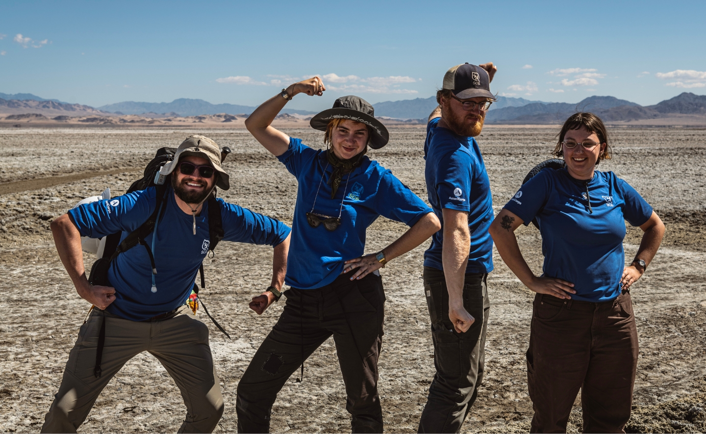 A group of four people wearing blue SCA t-shirts standing in a desert environment with mountains visible in the background. They are posing playfully, flexing their arms and striking confident stances. Each person is wearing outdoor gear, including hats and backpacks, suited for the rugged and arid terrain.