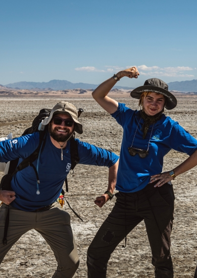 Two people wearing blue SCA t-shirts standing in a desert environment with mountains visible in the background. They are posing playfully, flexing their arms and striking confident stances. Each person is wearing outdoor gear, including hats and backpacks, suited for the rugged and arid terrain.