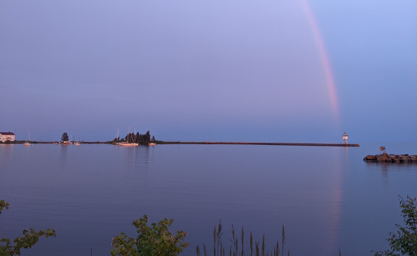 Panoramic view of a tranquil harbor of Lake Superior at dusk, featuring a lighthouse at the end of a long pier and a faint rainbow in the sky leading directly down to it.