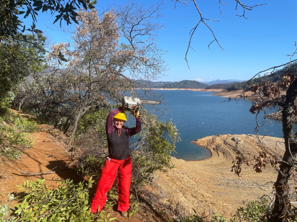 Kelly Graner in a yellow hard hat and red pants raises a chainsaw over head, working along a trail by a large body of water under a clear sky in Shasta, California.