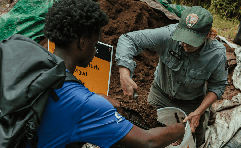 A person in a green uniform filling an SCA crew member's bucket with dirt