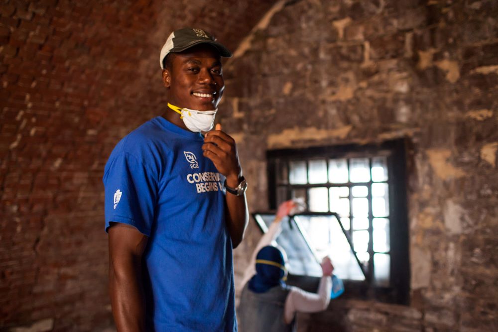 An SCA Historic Preservation Corps member wearing a "Conservation Begins Here" t-shirt and a cap, smiling inside a rustic brick building. The person is holding a face mask down around their chin.