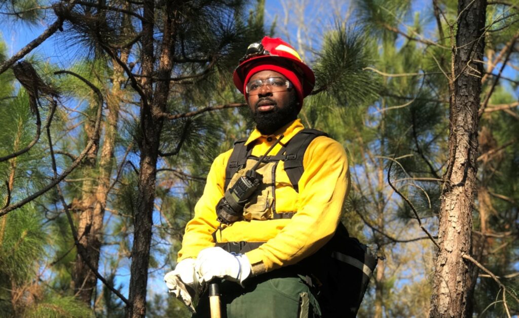 SCA Alum Eric Lucas stands in front of tall fir trees in forest. He wears fire service safety equipment including a red helmet.