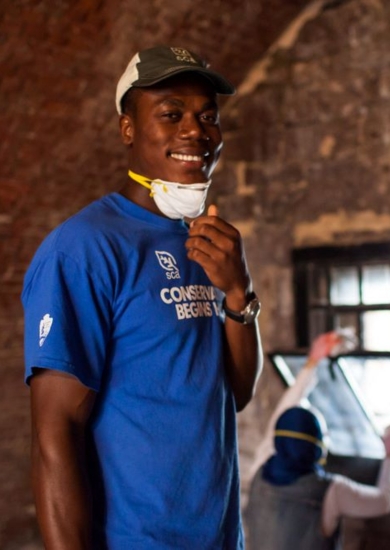 An SCA Historic Preservation Corps member wearing a "Conservation Begins Here" t-shirt and a cap, smiling inside a rustic brick building. The person is holding a face mask down around their chin.