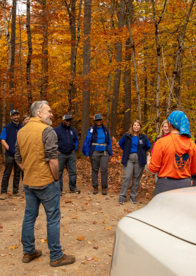 A circle of people listen to a young person where a bright orange shirt and blue bandana. They stand in a wooded area in Bear Brook State Park, New Hampshire.