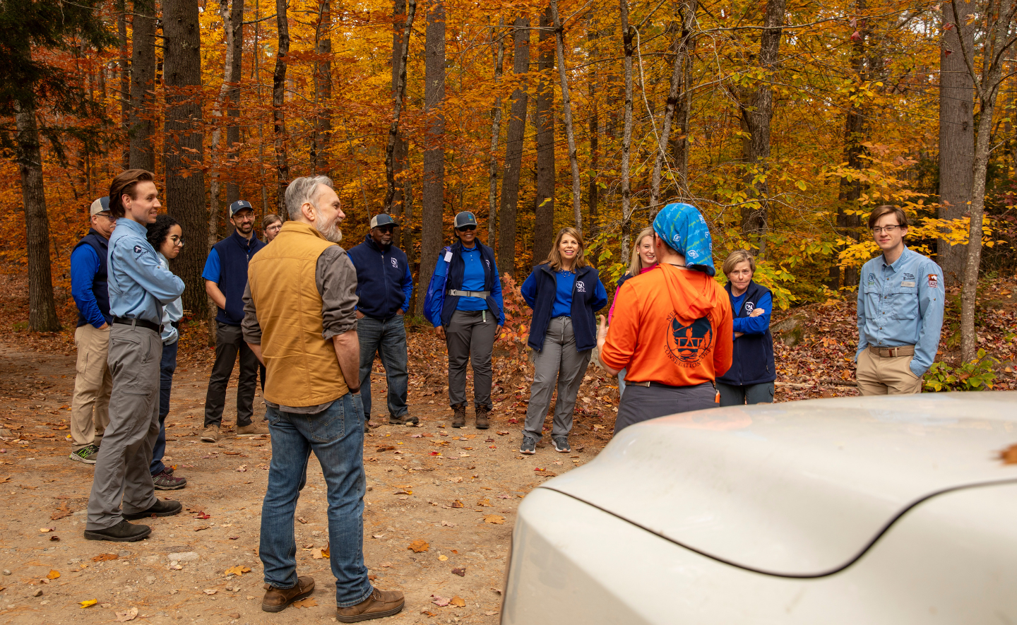 A circle of people listen to a young person where a bright orange shirt and blue bandana. They stand in a wooded area in Bear Brook State Park, New Hampshire.