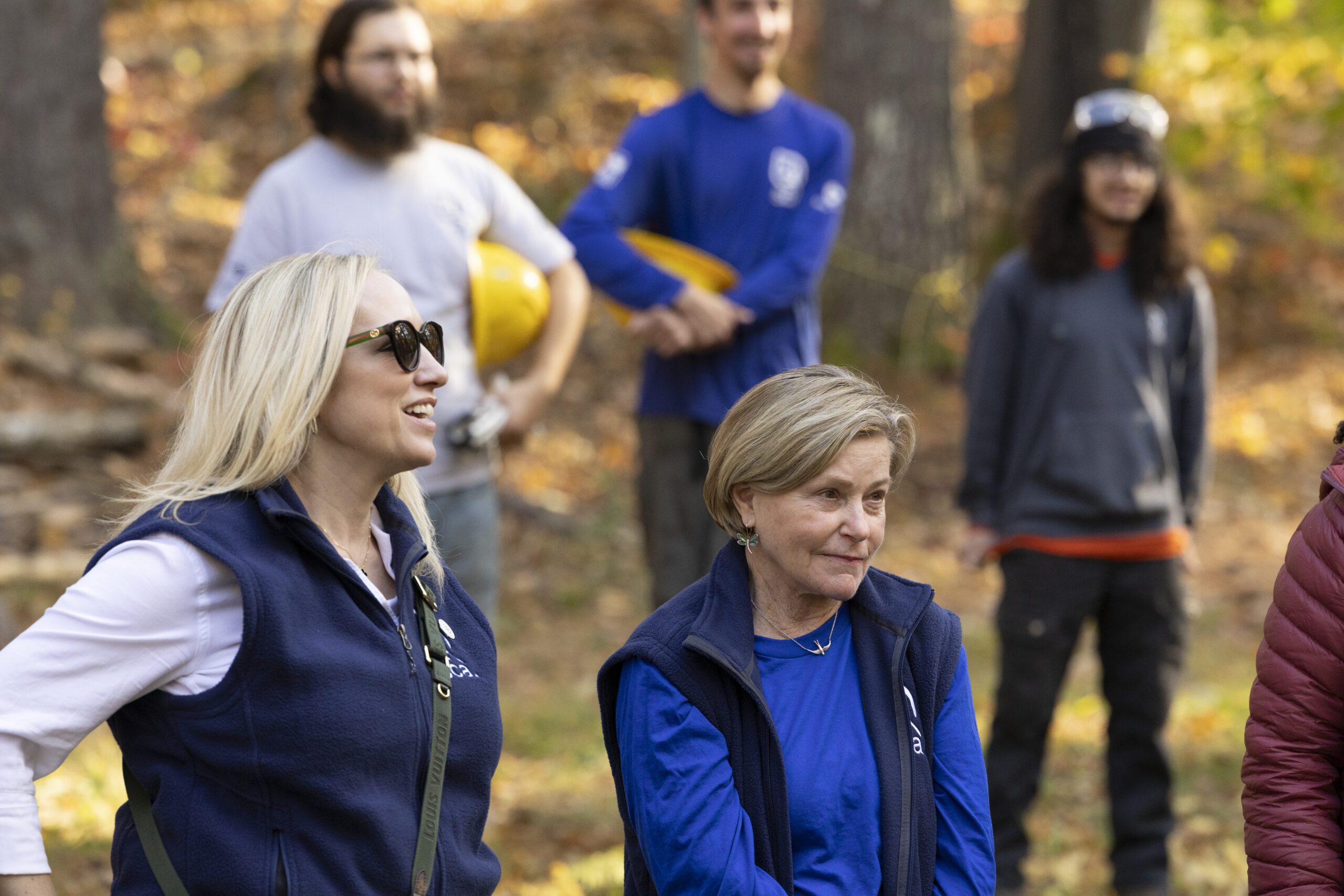 Two people wearing SCA branded clothing stand listening to a speaker at Bear Brook State Park, NH.