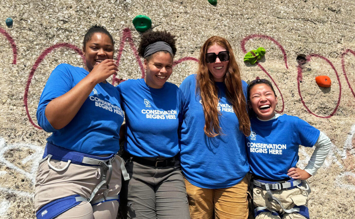 Four members of the 2024 Chicago Women Crew wearing blue SCA shirts stand together, smiling, in front of a rock-climbing wall. Two of the women wear climbing harnesses. 