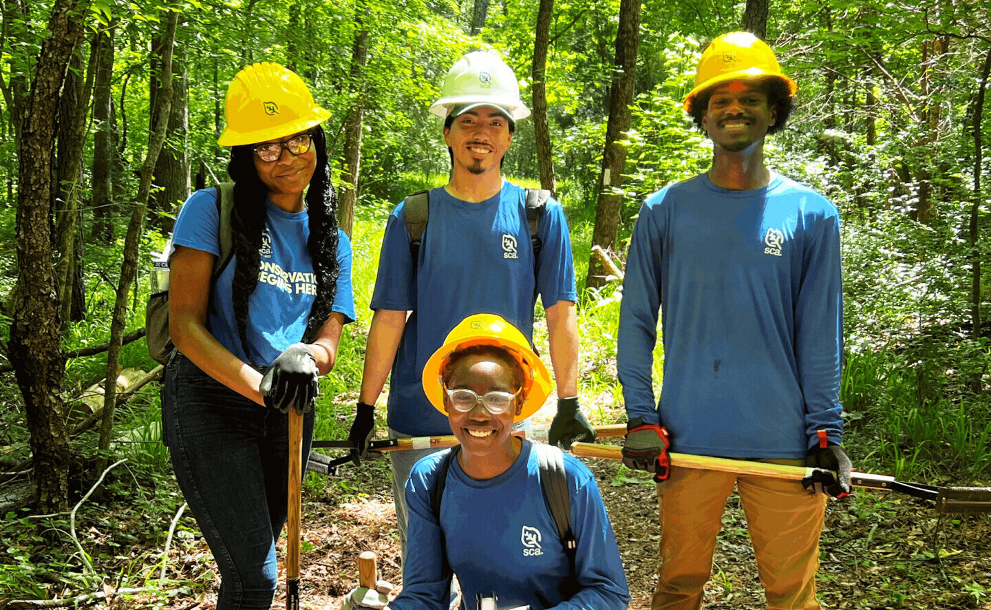 Four crew members from the SCA Atlanta UPS Summer Crew, all wearing blue shirts and hard hats, stand with work tools on a trail in a wooded area.