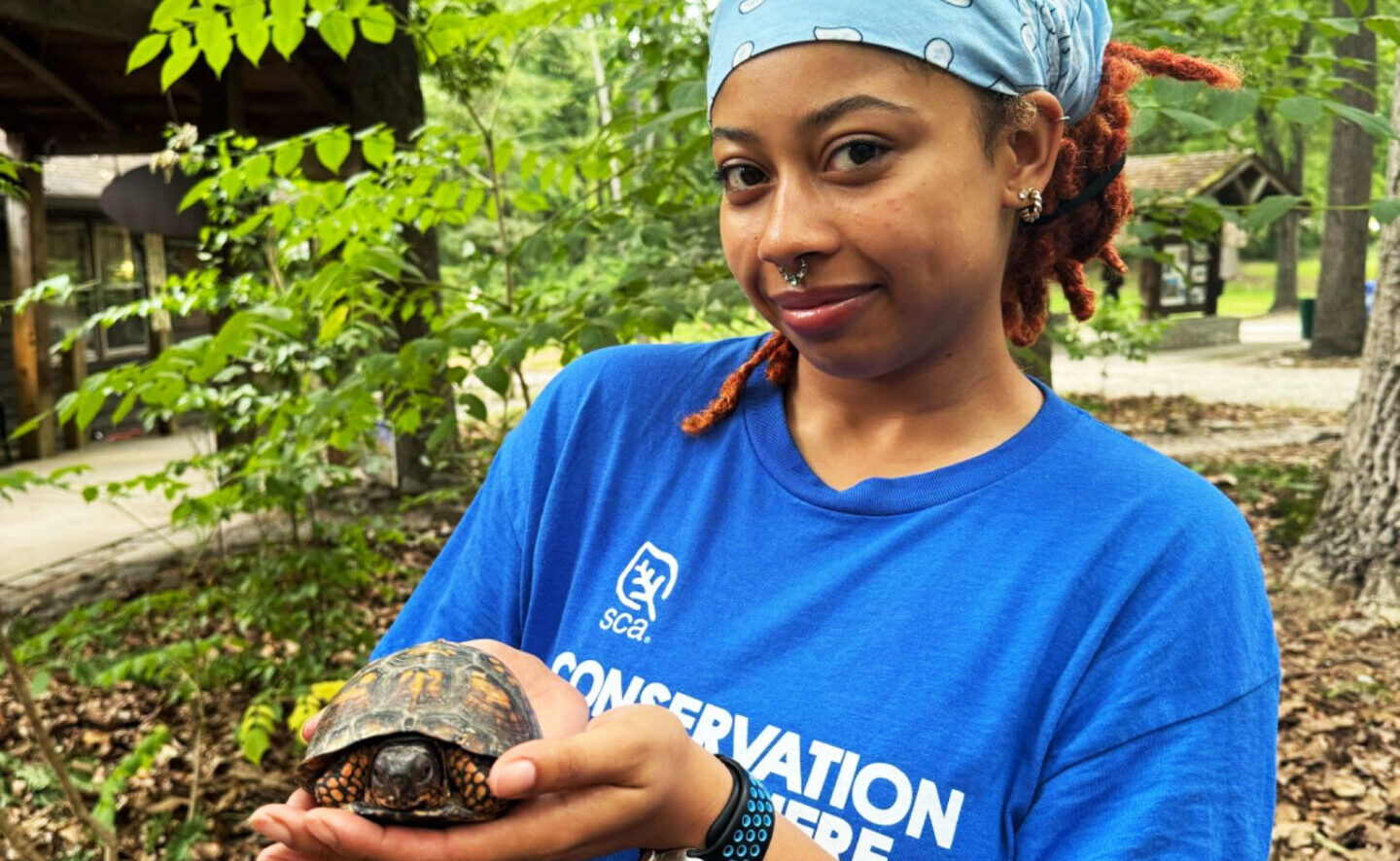 A young Black woman in a bright blue SCA shirt holds up a box turtle. 