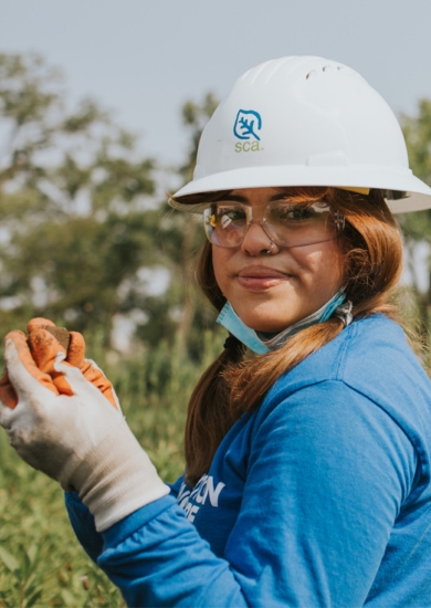 An SCA crew member with long reddish pigtails, wearing a long-sleeve blue SCA shirt, white hard hat and gardening gloves stands in a field of tall flowering plants.