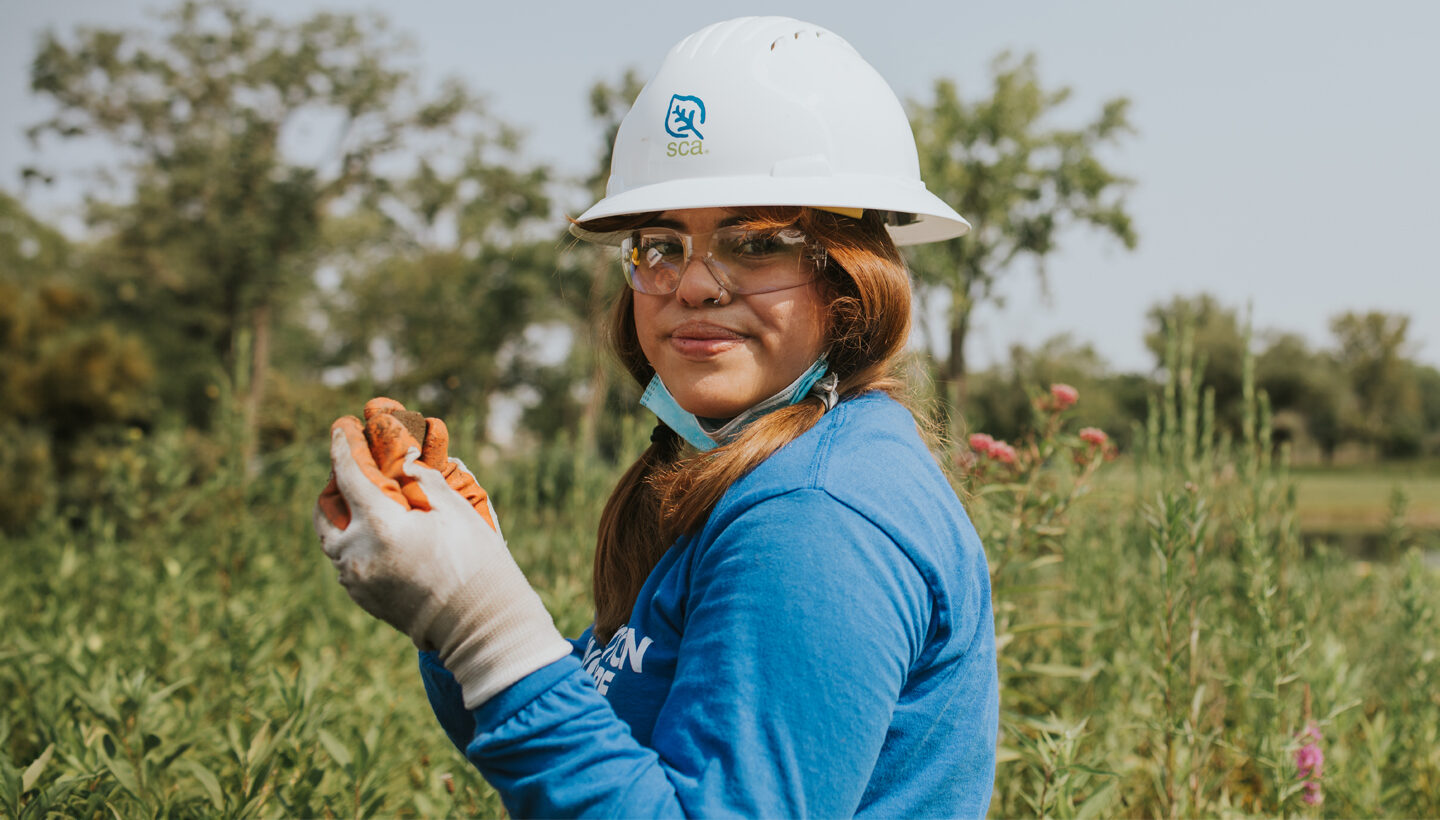 An SCA crew member with long reddish pigtails, wearing a long-sleeve blue SCA shirt, white hard hat and gardening gloves stands in a field of tall flowering plants.