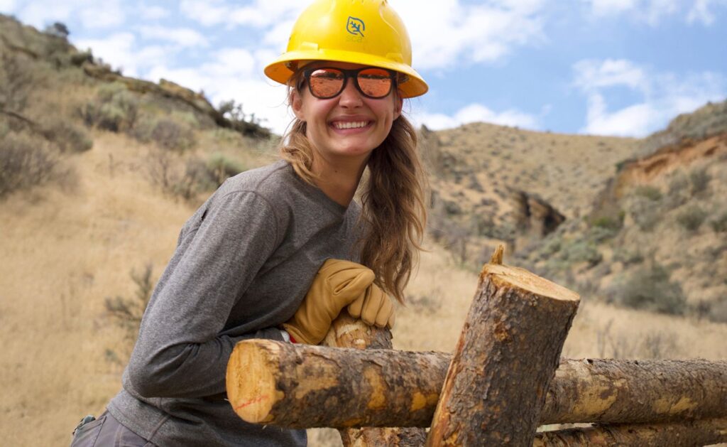 SCA crew member wearing a hard hat and gloves leaning on a wood fence