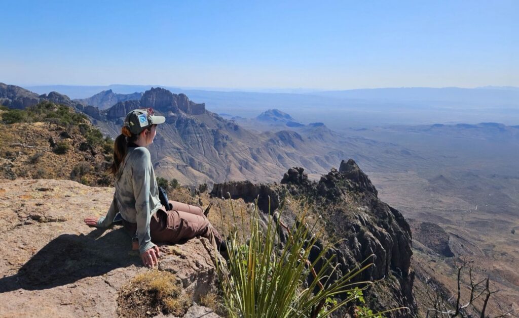 Photograph of an SCA crew member sitting on a rocky cliff at Big Bend National Park in Texas overlooking a vast mountainous desert landscape under a clear blue sky. The scene highlights rugged rock formations, sparse vegetation, and expansive distant valleys, conveying a sense of tranquility and natural beauty.