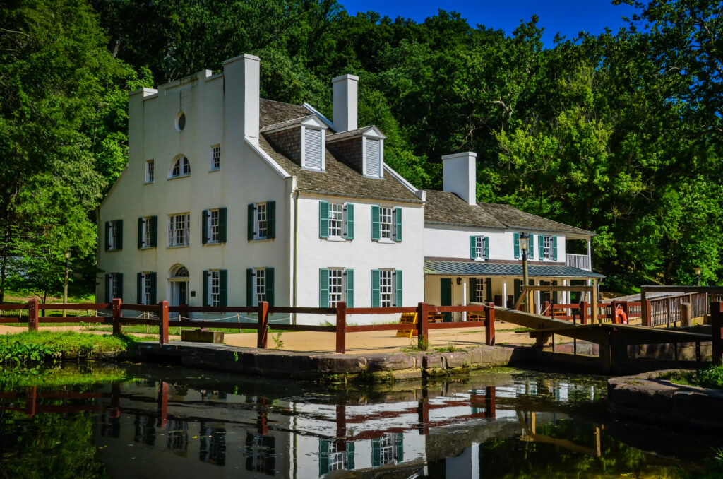 Photograph of the historic C&O Canal National Historical Park Visitor Center. The white building with green shutters is situated beside a calm canal, surrounded by lush green trees and features multiple chimneys, a wooden fence, and a small bridge crossing the canal, reflecting a serene and picturesque setting.