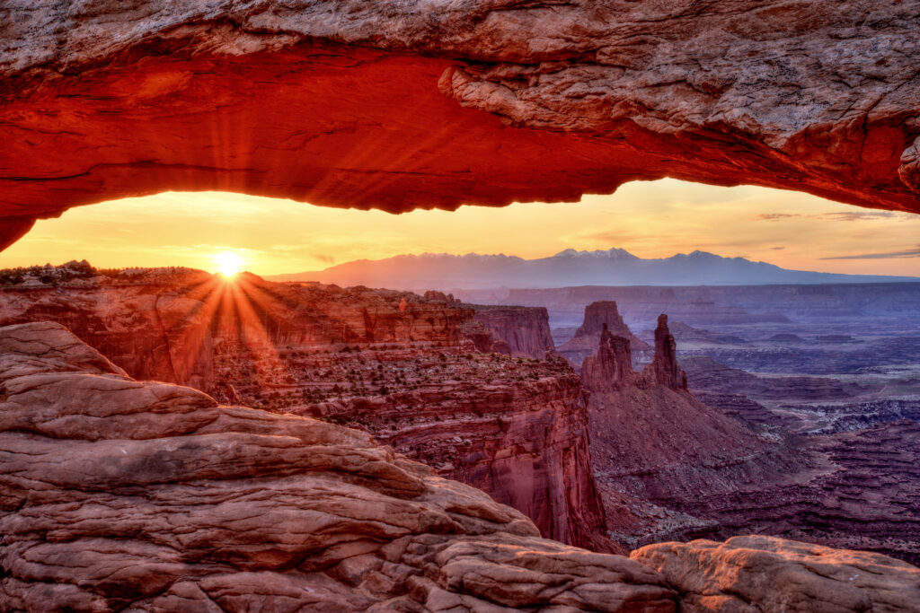 A photo of Mesa Arch at Sunrise at Canyonlands National Park in Utah shows a natural rock arch framing a sunrise over a vast canyon landscape with layered red rock formations and distant mountains. Warm orange and yellow sunlight contrasts with deep shadows, highlighting textures of the arch and rugged terrain below.