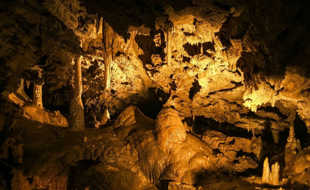 Photograph of a cave interior at Oregon Caves National Monument Preserve showcasing stalactites hanging from the ceiling and stalagmites rising from the ground, illuminated by warm yellow lighting. The image highlights natural rock formations with varied textures and shapes, emphasizing geological features typical of limestone caves.