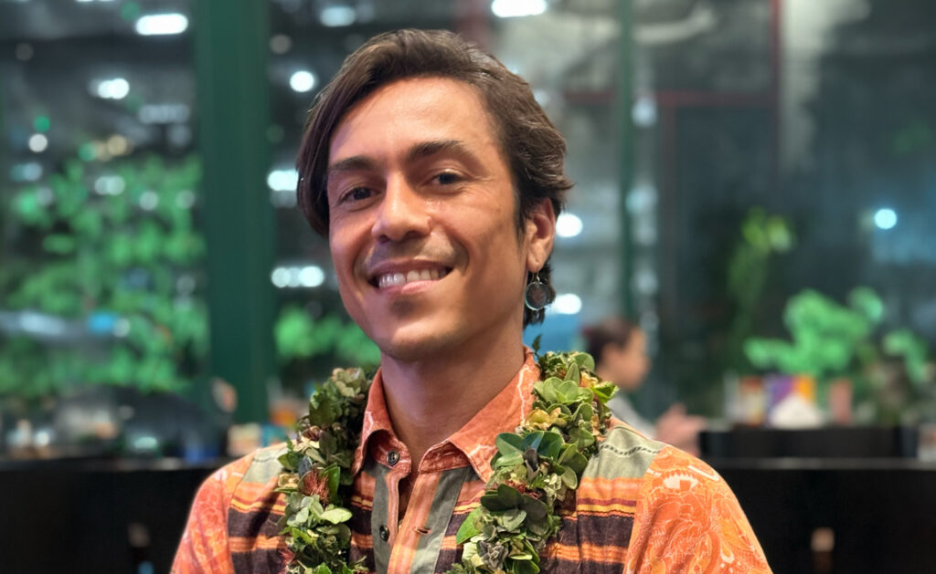 Photograph of a smiling man wearing an orange patterned shirt and a green leafy lei around his neck, set against a blurred background with plants and lights. The image captures a casual, warm atmosphere with natural tones and soft lighting highlighting the man's face.