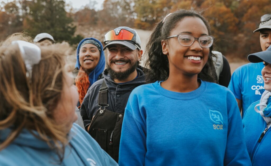 Photograph shows a diverse group of SCA crew members smiling outdoors, including people wearing blue shirts with an SCA logo. Autumn trees and casual work gear suggest a community conservation or service event, with participants gathered for teamwork and connection.