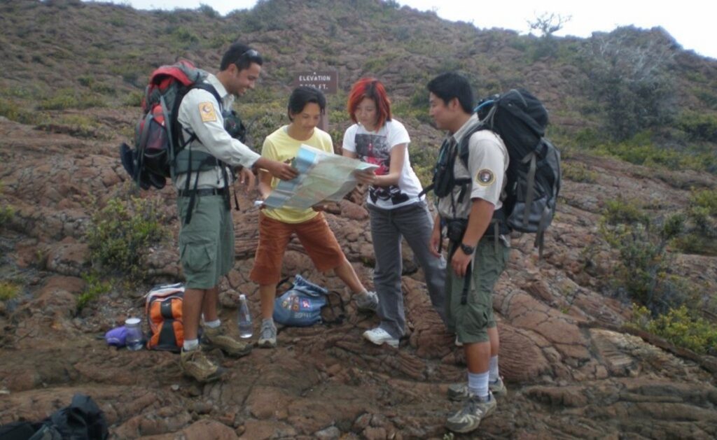 Photograph of two park rangers helping two hikers with a map on rugged volcanic terrain during an outdoor expedition. Backpacks, hiking gear, water bottles, sparse shrubs, and rocky hills surround group, emphasizing navigation and field exploration.