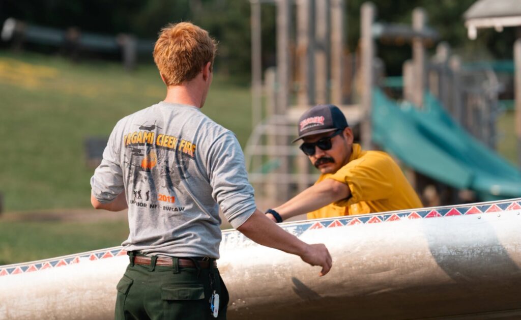 Photograph shows two men moving a canoe, with one wearing a yellow shirt, sunglasses, and mustache. A grassy venue, wooden structures, and teal slide appear in background, while patterned red-and-blue trim decorates canoe.