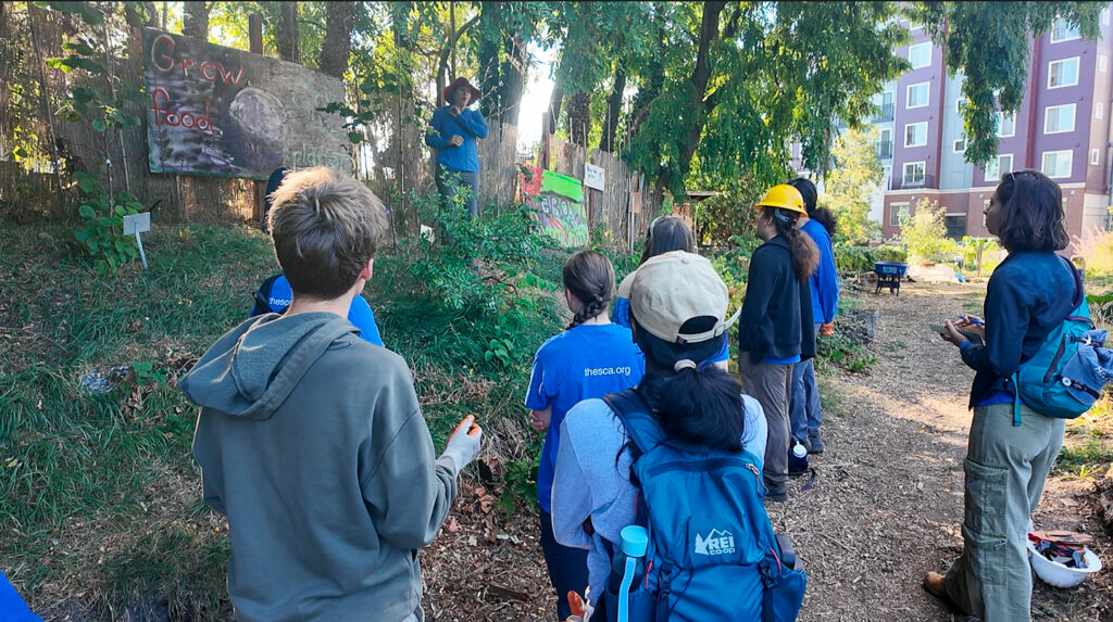 Photograph of the Seattle Urban Green team exploring an urban community garden with a guide. Crew members gather near a fence with handmade signs with apartment buildings visible behind trees.