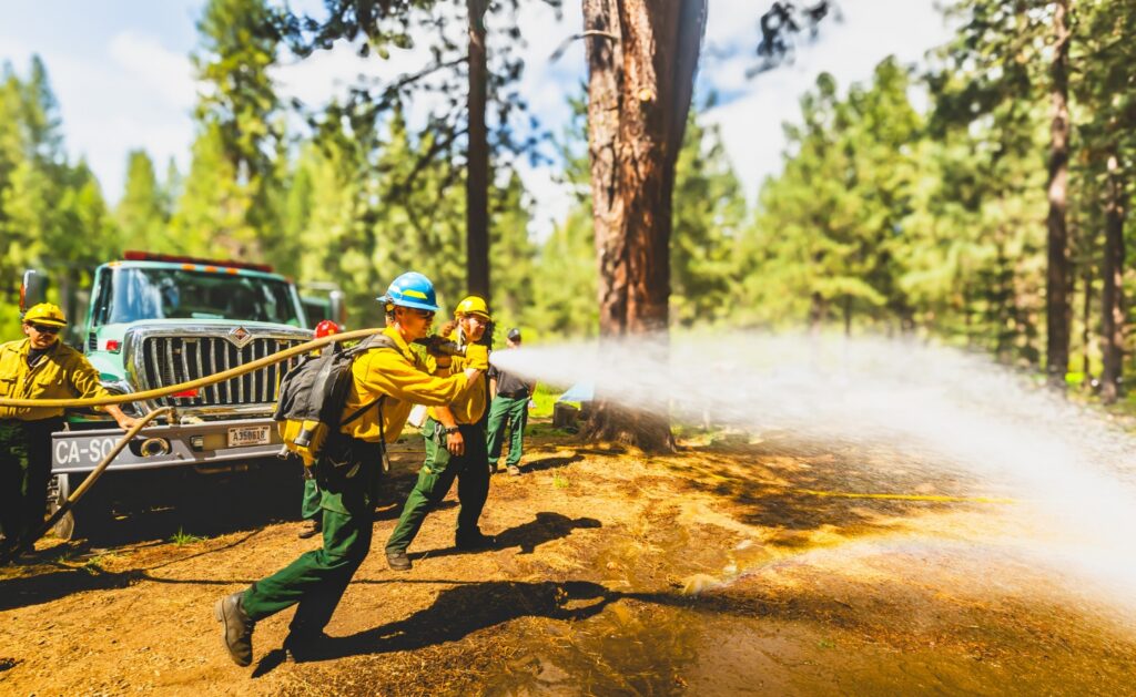 Photograph shows SCA Wildland Fire Academy cadet in yellow protective gear spraying water from a hose beside a fire truck in a forest. Dense trees, dry ground, water mist, and coordinated crew response highlight active wildfire suppression.