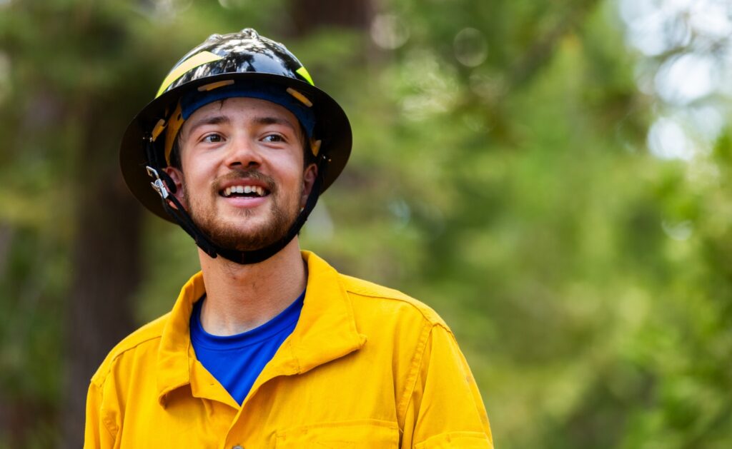 Photograph of smiling male SCA Wildland Fire Academy member wearing a black helmet and bright yellow protective jacket outdoors. Green forest background and blue undershirt create strong color contrast, highlighting a cheerful emergency responder during an outdoor assignment.